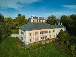 Widok z lotu ptaka na Pałac Ratibořice, otoczony ścieżkami i zielenią rozległego parku krajobrazowego. Budynek ma dach w kształcie litery T i leży wśród drzew o jesiennych barwach.