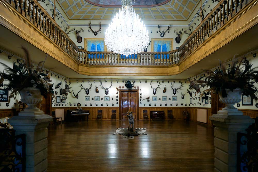 Reprezentacyjna sala myśliwska w Zamku Dětenice z galerią na piętrze, ozdobiona porożami i trofeami zwierząt. W centrum stoi rzeźba, a nad nią wisi ogromny kryształowy żyrandol.