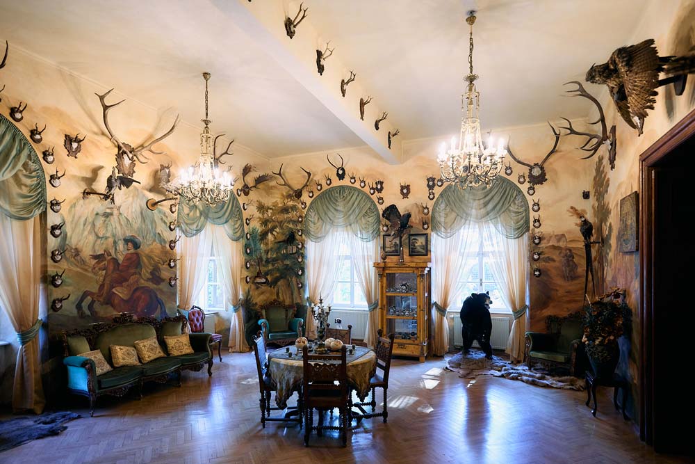 Myśliwski salon w Zamku Dětenice z malowidłami ściennymi przedstawiającymi sceny polowań, trofeami zwierząt i eleganckimi meblami. Wnętrze oświetlają dwa żyrandole.