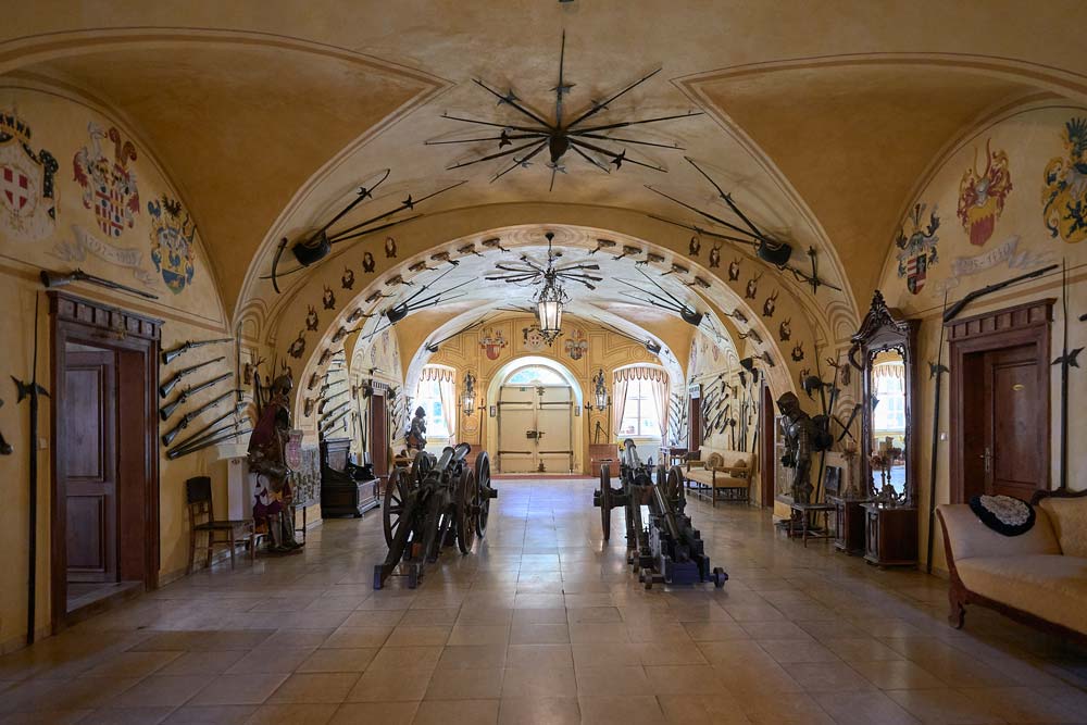 Zbrojownia w Zamku Dětenice z kolekcją dawnych broni i zbroi. Na ścianach wiszą halabardy, miecze i herby rodowe, a w centrum stoją armaty na drewnianych kołach.