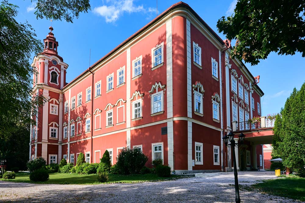 Barokowy Zamek Dětenice w intensywnie czerwonym kolorze z białymi detalami i narożną wieżą. Przed budynkiem znajduje się żwirowa droga i zieleń parku.