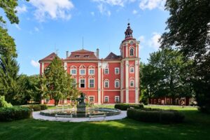 Główna fasada Zamku Dětenice z symetrycznym układem okien i balkonem wspartym na kolumnach. Na pierwszym planie widoczne są krzewy i rabata z czerwonych kwiatów.