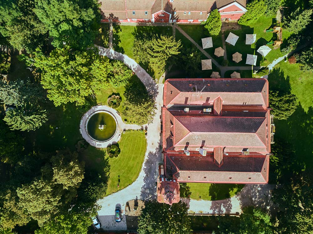 Ujęcie z drona na Zamek Dětenice z czerwonym dachem, otoczony parkiem z fontanną i alejkami. Widoczne są też ogrodowe altany i parasole.