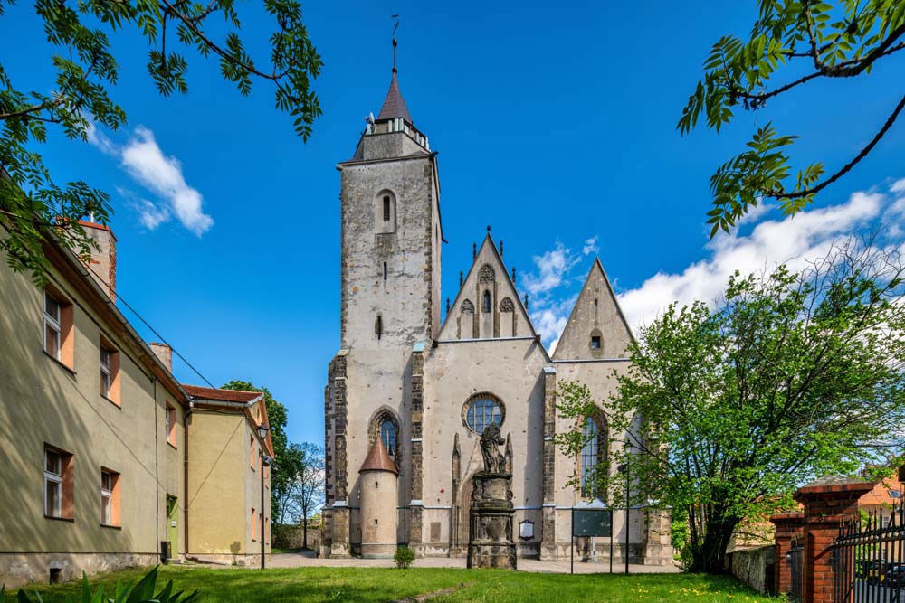Gotycki kościół św. Marcina w Jaworze z wysoką wieżą i dwoma smukłymi szczytami. Przed świątynią stoi barokowa figura, a otaczają ją drzewa i kamienice.