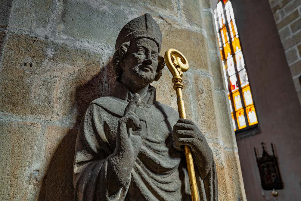 Kamienna rzeźba biskupa z pastorałem wewnątrz kościoła św. Marcina w Jaworze. Postać stoi przy filarze z kamienia, w tle widoczne są witraże.