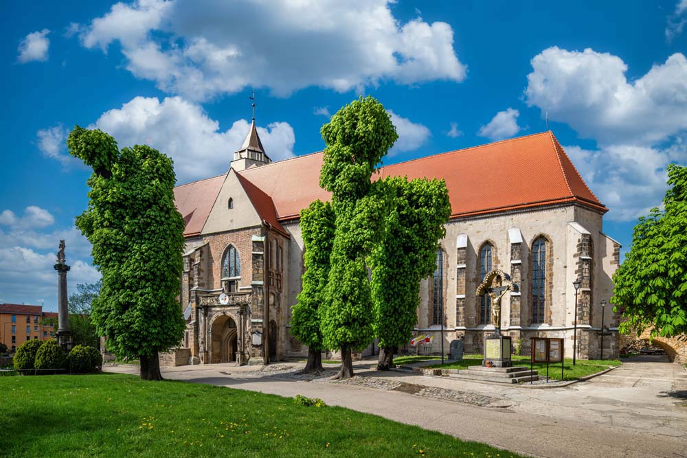 Widok na kościół św. Marcina w Jaworze z czerwonym dachem i gotyckimi oknami. W otoczeniu świątyni rosną przystrzyżone drzewa, a na placu stoi kamienny krzyż.