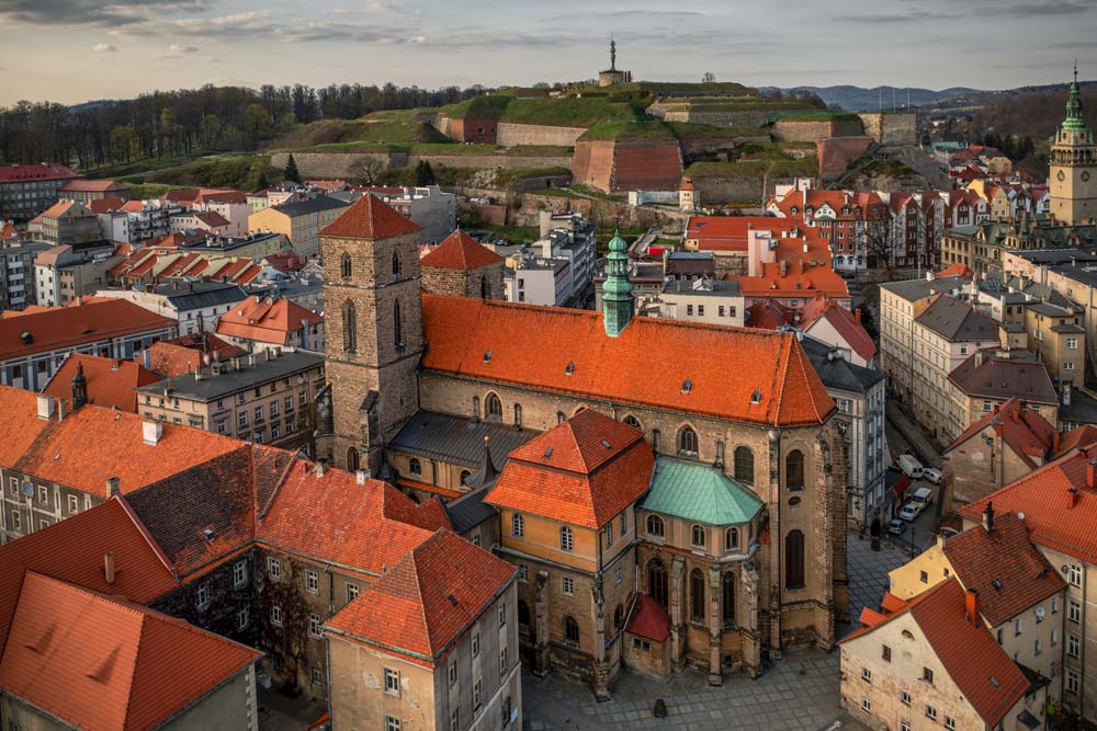 Kościół Najświętszej Maryi Panny w Kłodzku z czerwonym dachem i kamienną wieżą, otoczony kamienicami Starego Miasta, z widoczną twierdzą na wzgórzu w tle.