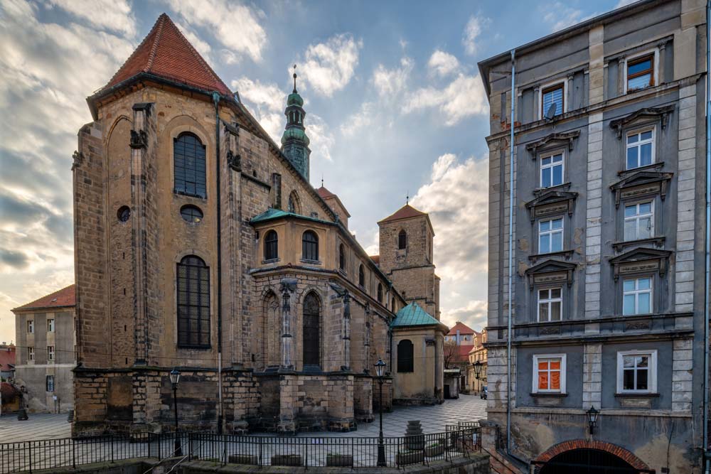 Kościół Wniebowzięcia Najświętszej Maryi Panny w Kłodzku widziany z placu, z wysokimi gotyckimi oknami i wieżą otoczoną miejską zabudową.