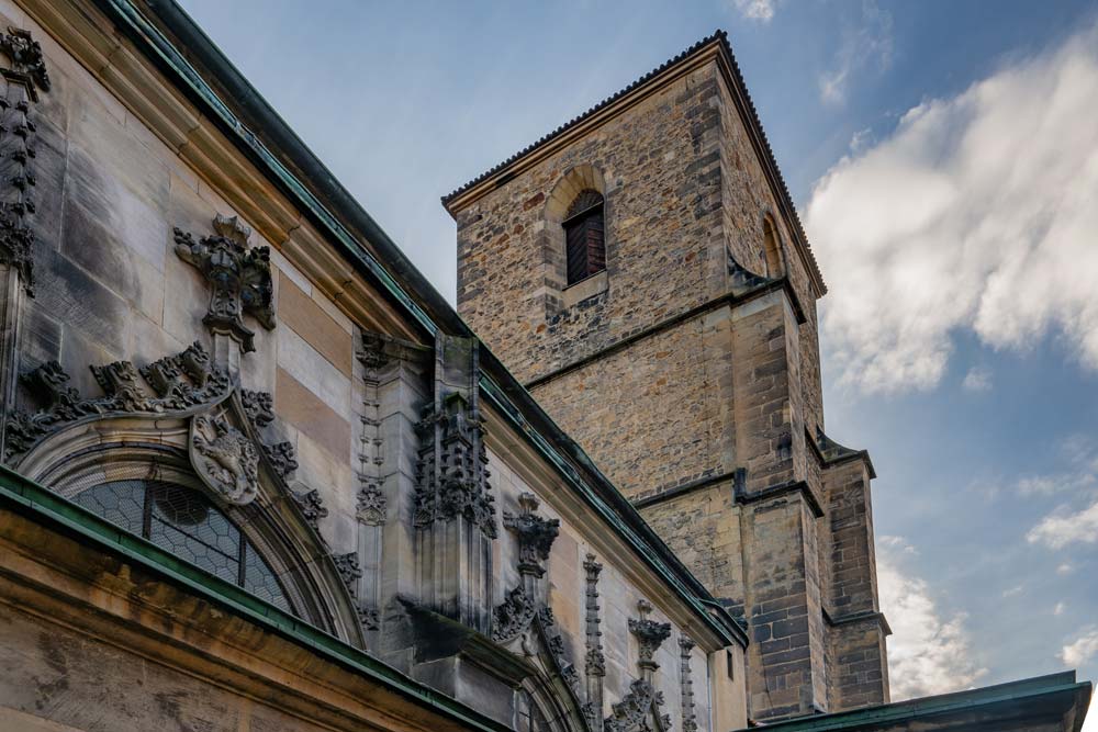 Zbliżenie na kamienną wieżę i fasadę Kościoła Wniebowzięcia Najświętszej Maryi Panny w Kłodzku z bogatymi barokowymi zdobieniami pod niebem z chmurami.