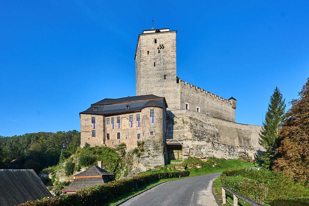 Zamek Kost widziany od strony drogi prowadzącej pod górę, z masywną kamienną wieżą i murami obronnymi wznoszącymi się nad zielonym wzgórzem w słoneczny dzień.