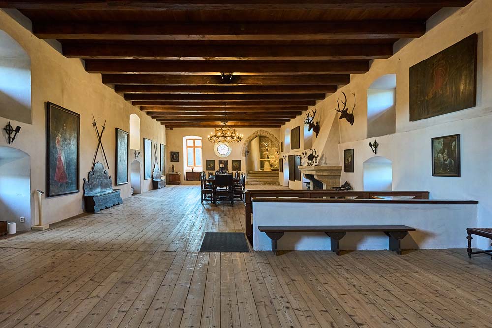 Wnętrze wielkiej sali Zamku Kost z drewnianym stropem, obrazami, trofeami myśliwskimi i długim stołem w centrum pomieszczenia.
