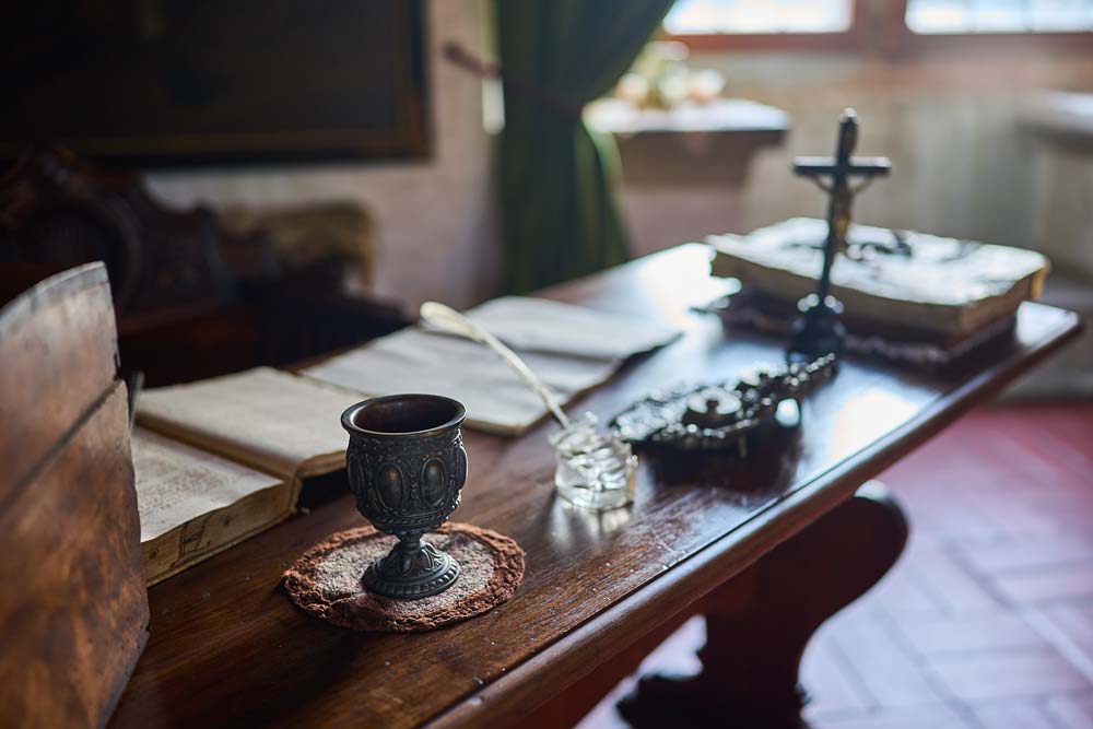 Zbliżenie na biurko w Zamku Kost z kielichem, krucyfiksem, otwartą księgą i piórem leżącymi na drewnianym blacie przy oknie.