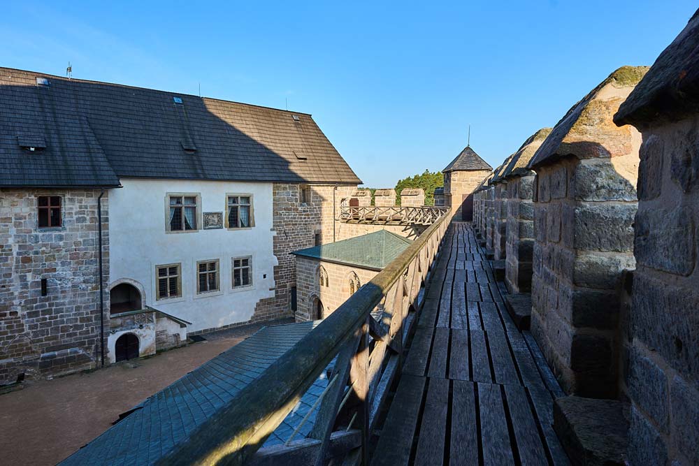 Widok z chodnika obronnego na dziedziniec Zamku Kost, z kamiennymi murami, wieżami i drewnianą balustradą biegnącą wzdłuż blanków.