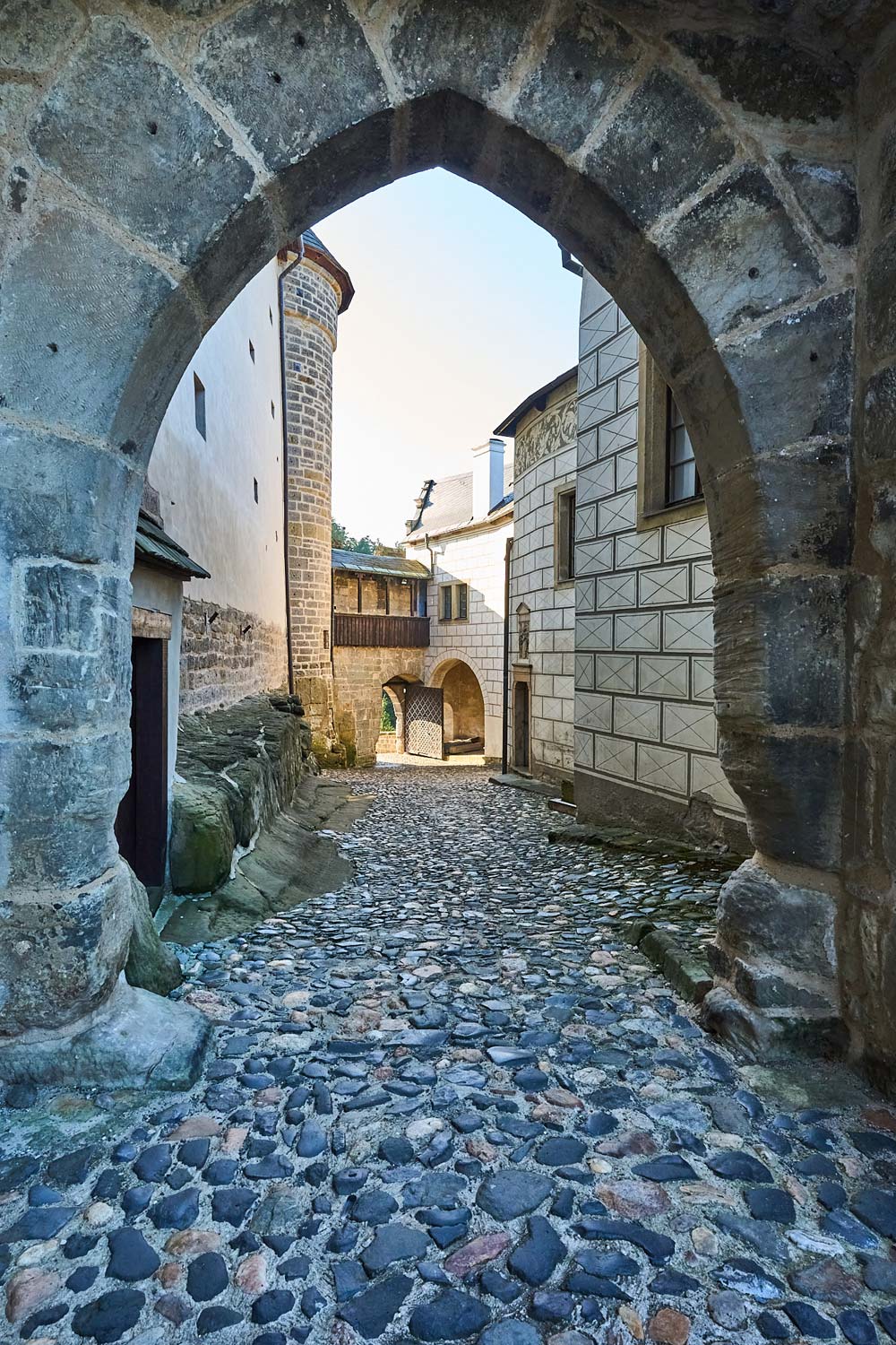 Kamienne przejście w Zamku Kost z brukowaną ścieżką prowadzącą przez łukowaty portal między średniowiecznymi murami zamkowymi.