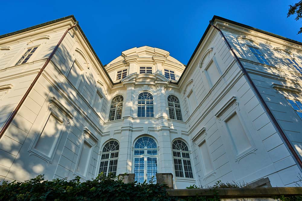 Zewnętrzny widok bocznej elewacji Zamku Karlova Koruna z wysokimi, łukowatymi oknami i białą barokową fasadą kontrastującą z błękitnym niebem.