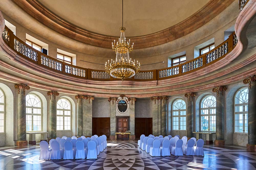 Okrągła sala balowa Zamku Karlova Koruna z marmurową posadzką w geometryczny wzór, dwoma kondygnacjami galerii i dużym złotym żyrandolem. Ustawiono tu krzesła na wydarzenie.