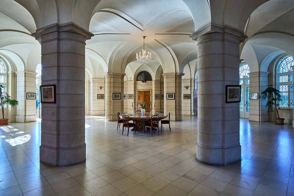 Przestronne wnętrze Zamku Karlova Koruna z kamiennymi kolumnami i dużymi oknami w półkolistych ramach. Centralny punkt stanowi stół otoczony krzesłami pod żyrandolem.