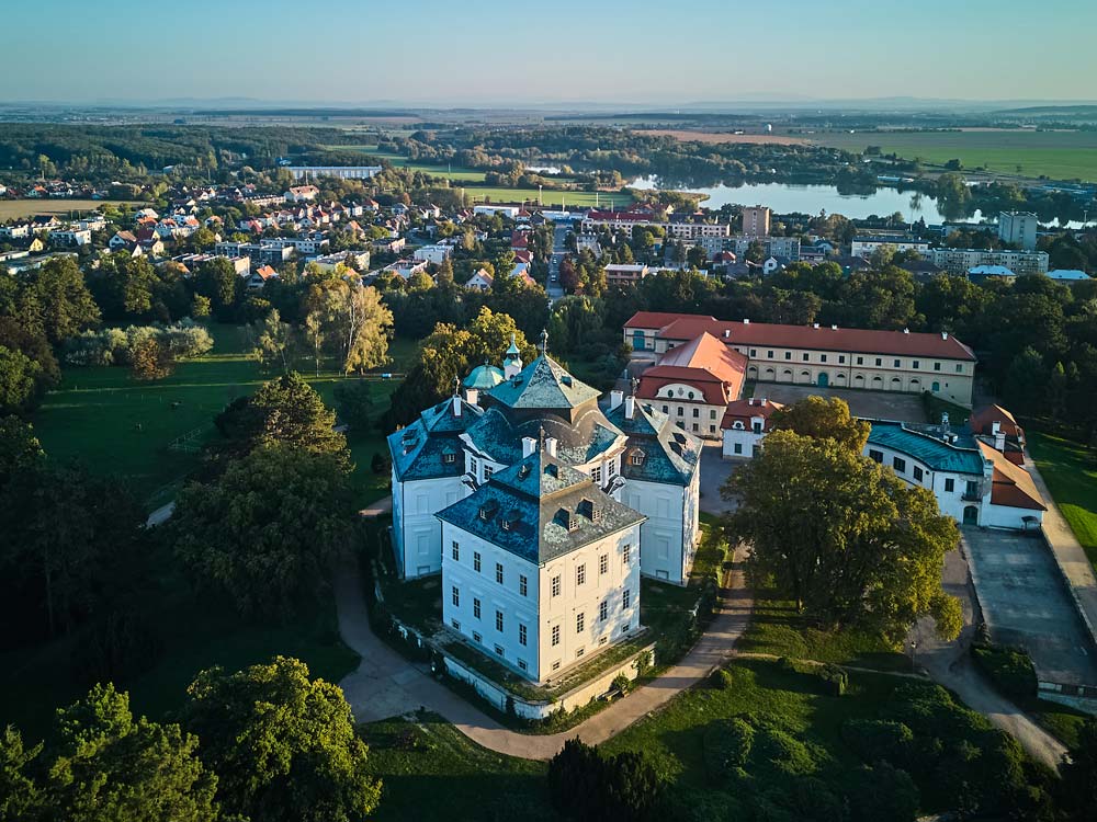 Ujęcie z drona na Zamek Karlova Koruna z otaczającym go parkiem, zabudowaniami folwarcznymi i panoramą miasta Chlumec nad Cidliną z jeziorami w tle.
