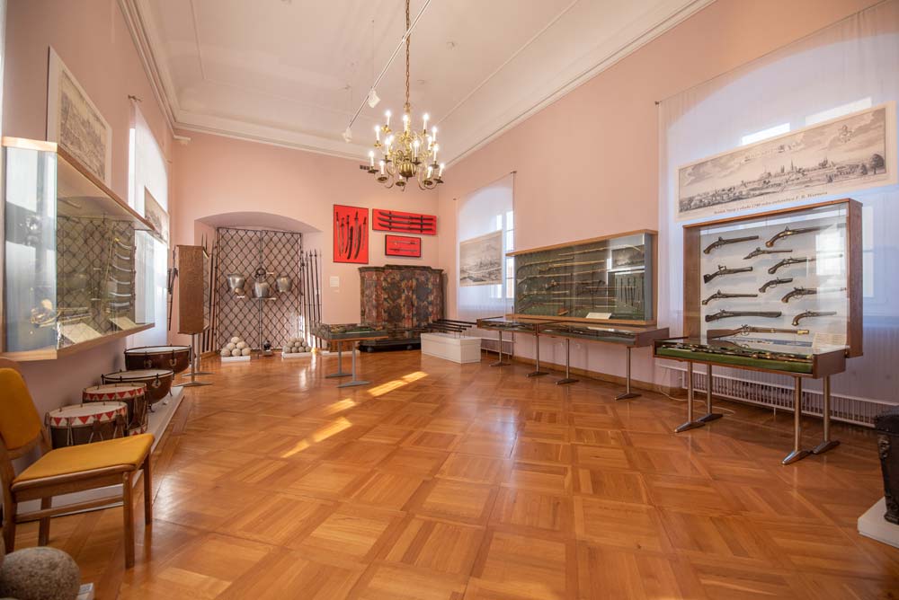 Wnętrze sali militariów w dawnym Pałacu Biskupim w Nysie, prezentujące zabytkowe bronie palne i białe w szklanych gablotach. Na ścianach wiszą ryciny i ozdobne elementy ekspozycji.