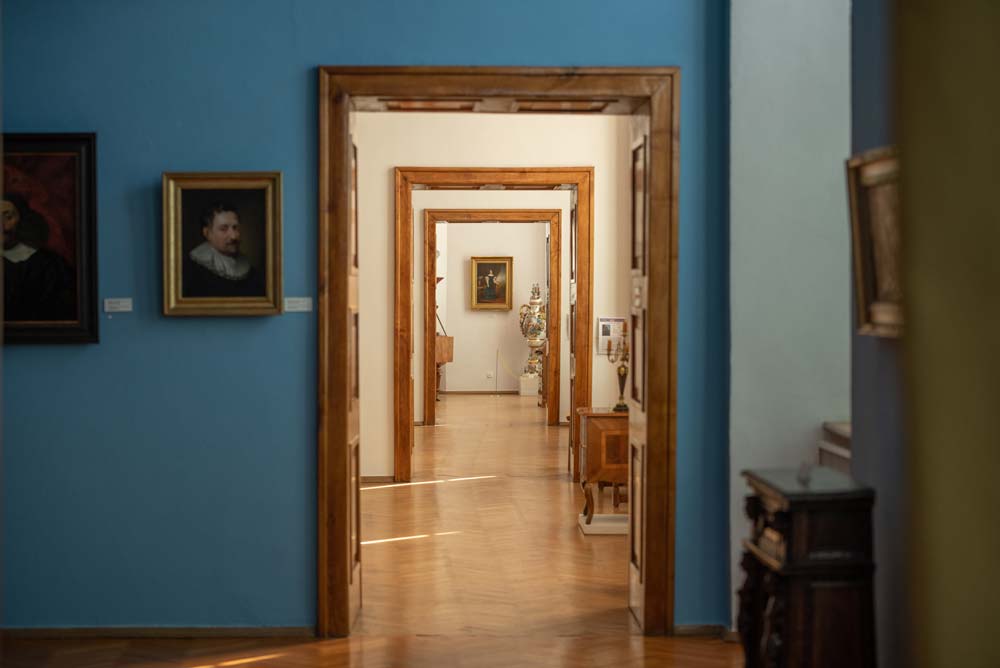 Widok przez kilka otwartych drzwi w dawnym Pałacu Biskupim w Nysie, ukazujący kolejne sale muzealne z obrazami, meblami i dekoracyjnym wazonem w głębi. Ściany w różnych kolorach tworzą efekt perspektywy.