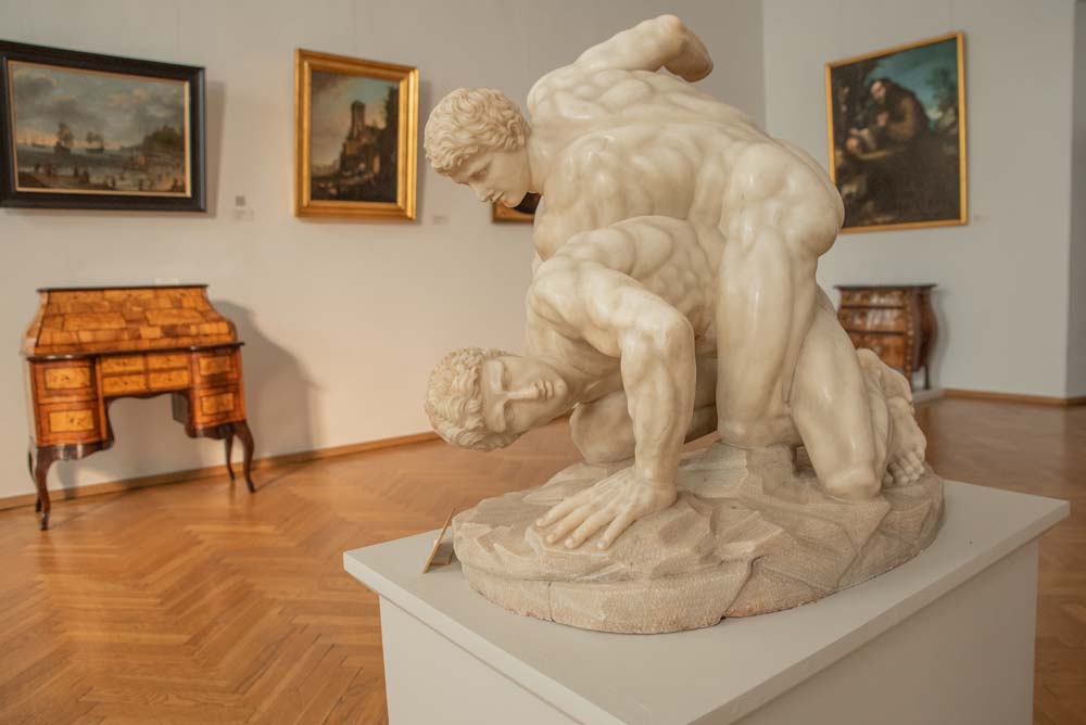 Ekspozycja w dawnym Pałacu Biskupim w Nysie, przedstawiająca marmurową rzeźbę dwóch zapaśników. W tle widoczne obrazy w złotych ramach i zabytkowe biurko.