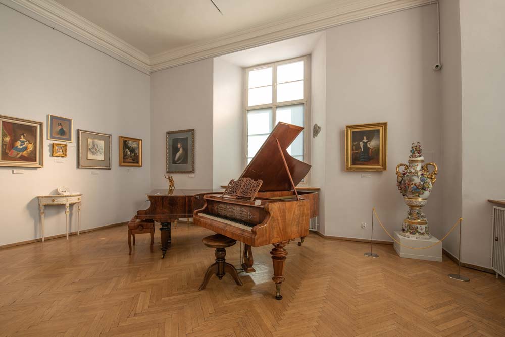 Wnętrze sali muzealnej w dawnym Pałacu Biskupim w Nysie, z dwoma drewnianymi fortepianami o otwartych pokrywach. Na ścianach wiszą klasyczne obrazy, a w rogu stoi duży porcelanowy wazon.