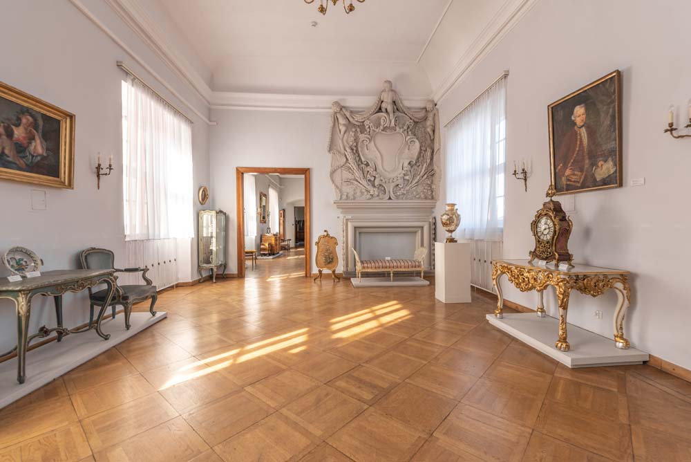 Jasna sala w dawnym Pałacu Biskupim w Nysie z ozdobnym barokowym kominkiem, zabytkowymi meblami i złoconym zegarem. Na ścianach wiszą obrazy, a przez duże okna wpada światło dzienne.