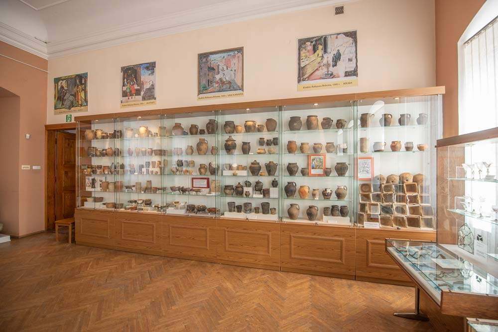 Sala archeologiczna w dawnym Pałacu Biskupim w Nysie, z przeszklonymi gablotami wypełnionymi glinianymi naczyniami i fragmentami ceramiki. Nad nimi wiszą ilustracje o tematyce archeologicznej.