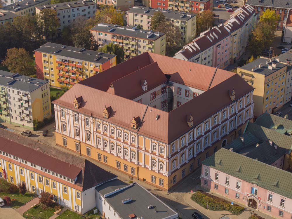 Ujęcie z lotu ptaka na barokowy pałac biskupi w Nysie z czerwoną dachówką i zdobioną fasadą. Budynek ma kształt prostokątnego dziedzińca i jest otoczony kolorowymi blokami mieszkalnymi.