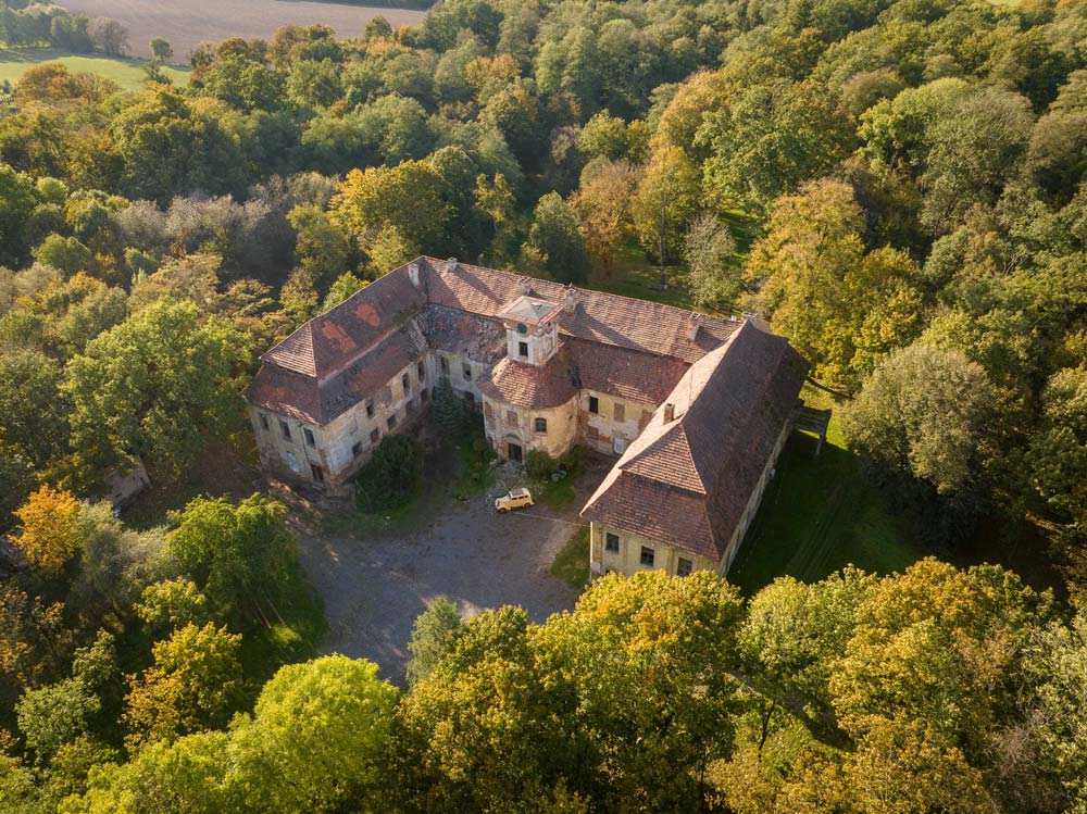 Widok z drona na Pałac Rozkochów otoczony barwnymi, jesiennymi drzewami. Ujęcie ukazuje cały kompleks z centralnym dziedzińcem i ceglanym dachem.