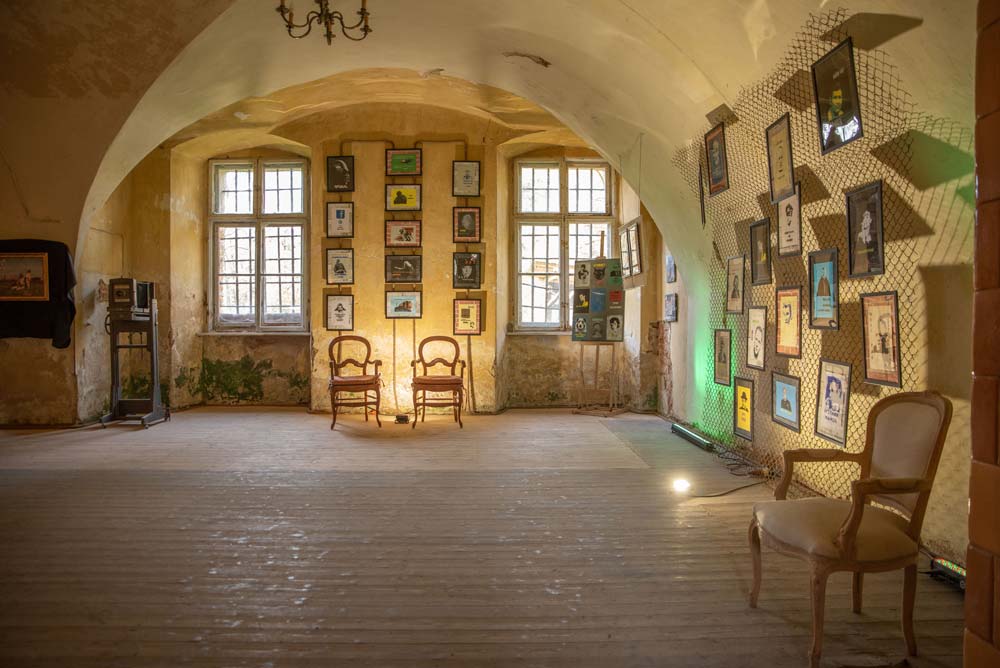 Wnętrze Pałacu Rozkochów z ekspozycją plakatów i fotografii artystycznych zawieszonych na ścianach o łukowych sklepieniach. Przy oknach stoją zabytkowe fotele.