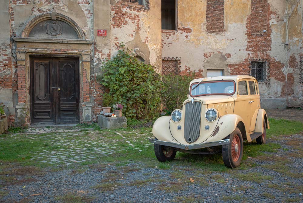Stary kremowy samochód z lat 30. XX wieku stoi przed zniszczoną fasadą Pałacu Rozkochów z odpadającym tynkiem i starymi drewnianymi drzwiami.