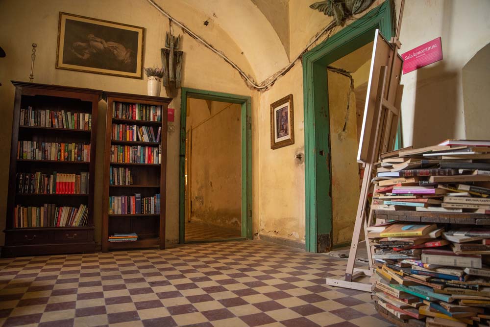 Pomieszczenie w Pałacu Rozkochów z regałami pełnymi książek, stosem woluminów na podłodze i starymi drzwiami w zielonych ramach prowadzącymi do kolejnych sal.