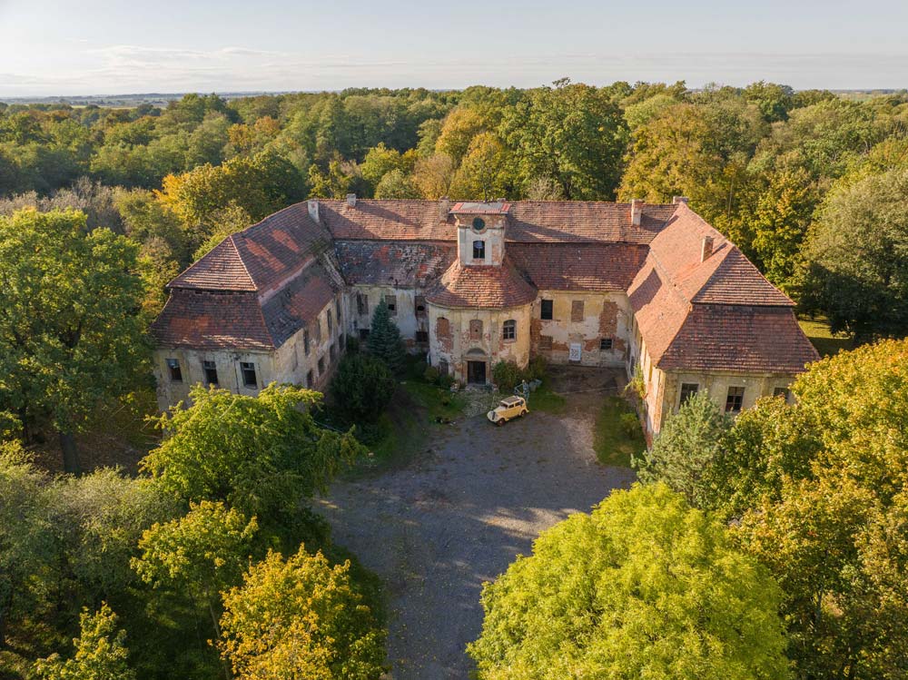 Widok z góry na Pałac Rozkochów położony pośród drzew. Ceglano-dachowa rezydencja tworzy zamknięty dziedziniec, w którego centrum stoi zabytkowy samochód.