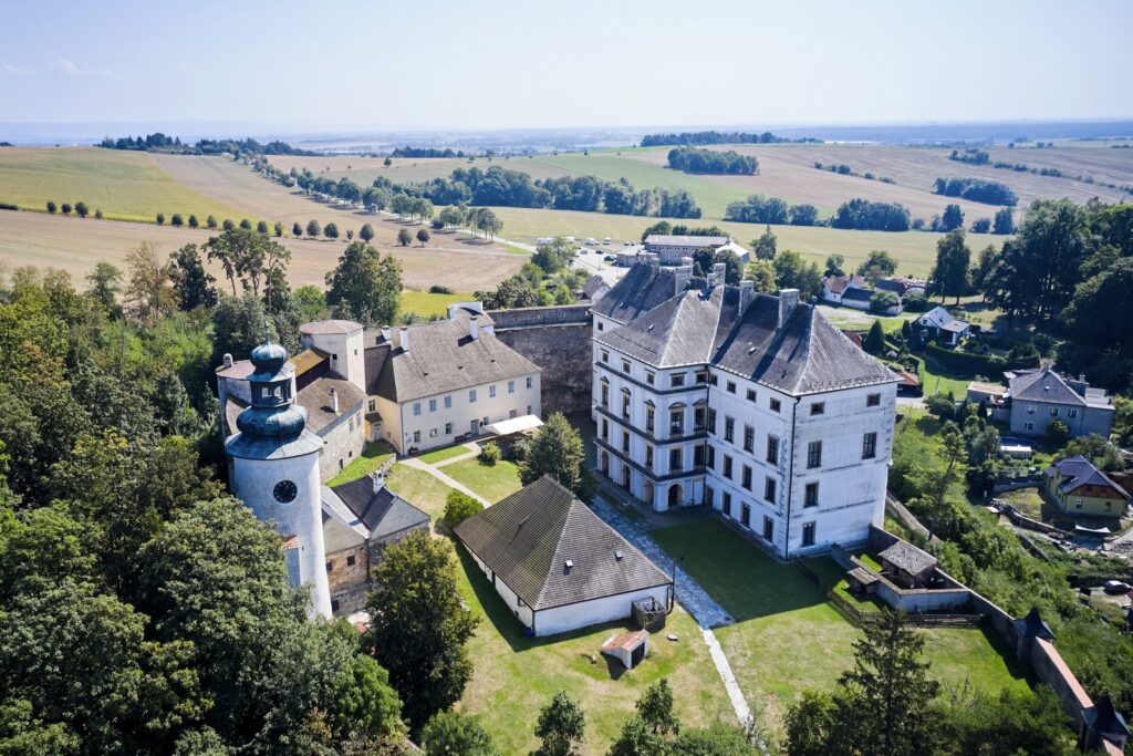 Widok z drona na Pałac Úsov na Morawach z charakterystyczną wieżą o cebulastym hełmie, dziedzińcem i otaczającymi go polami uprawnymi.