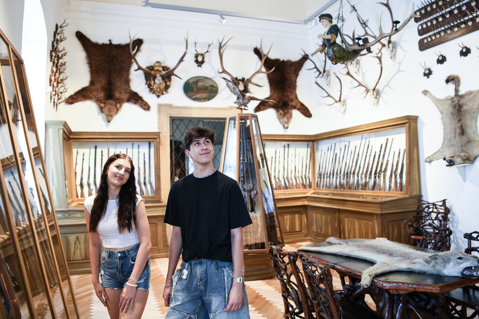 Wystawa dawnych strzelb, trofeów i skór zwierząt w Pałacu Úsov. Dwoje zwiedzających ogląda bogatą kolekcję eksponatów zgromadzonych w drewnianych gablotach.