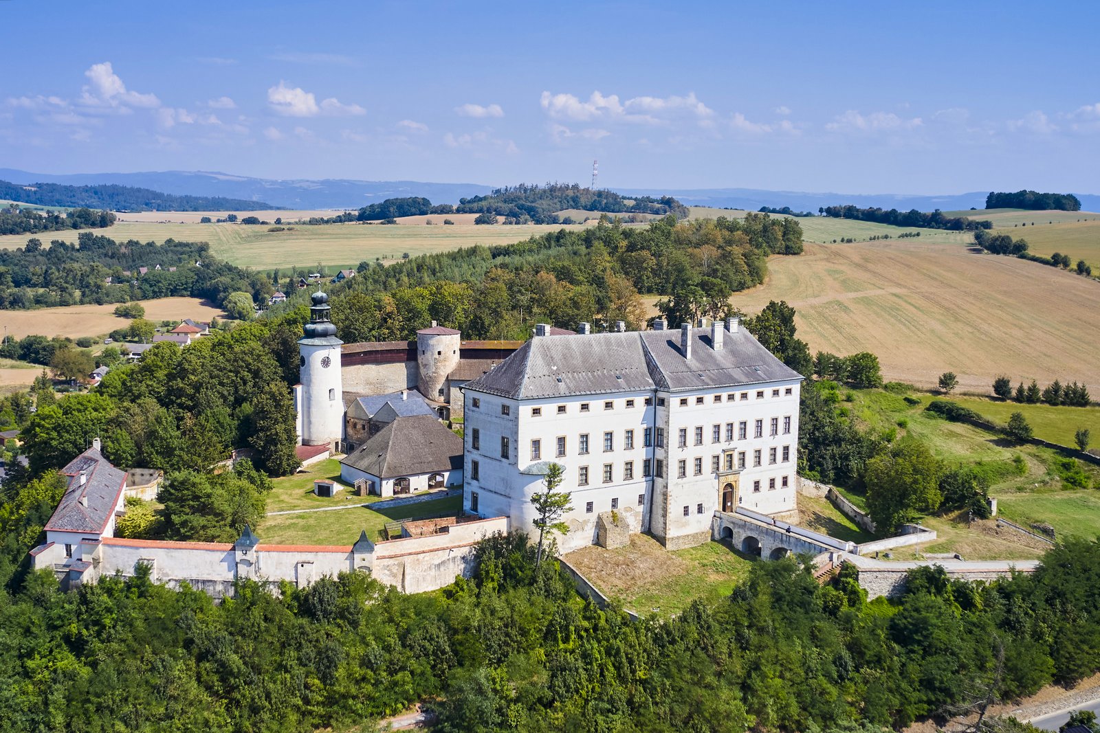 Zamek i pałac w Úsovie sfotografowany z drona. Kompleks otoczony jest murami obronnymi, z charakterystyczną wieżą zegarową i budynkami gospodarczymi pośród pól.