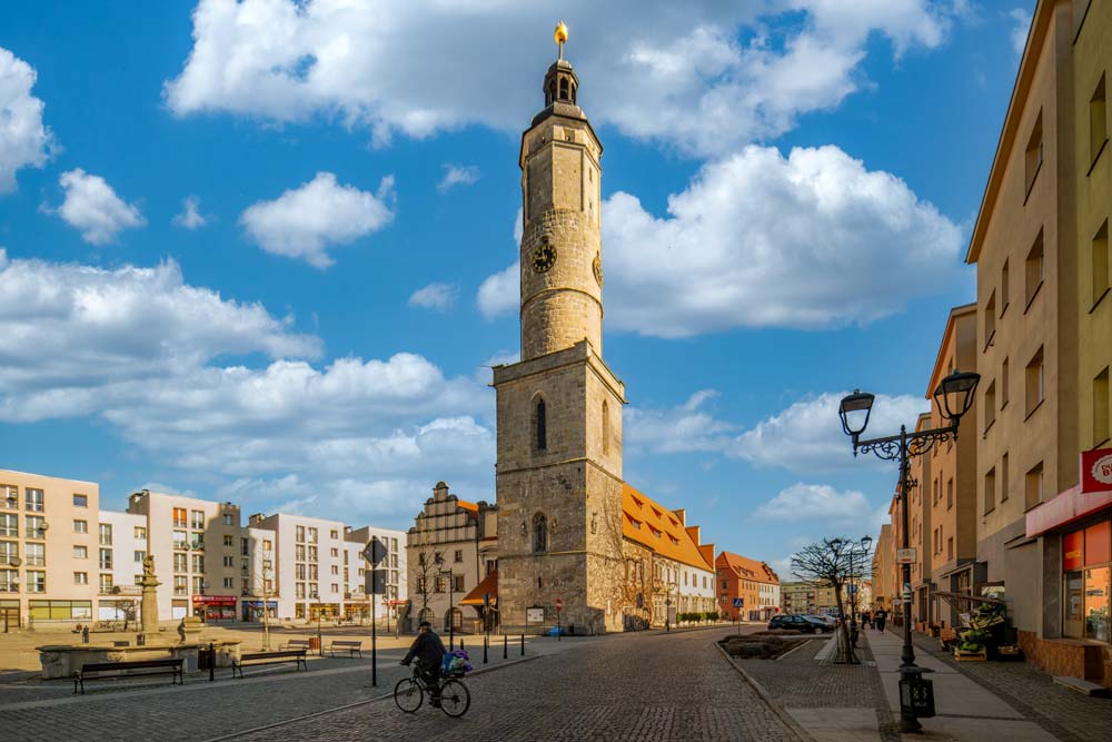 Wieża Ratusza w Lwówku Śląskim z zegarem i czerwonymi dachami budynków rynkowych. Na ulicy widać przechodniów i rowerzystę.
