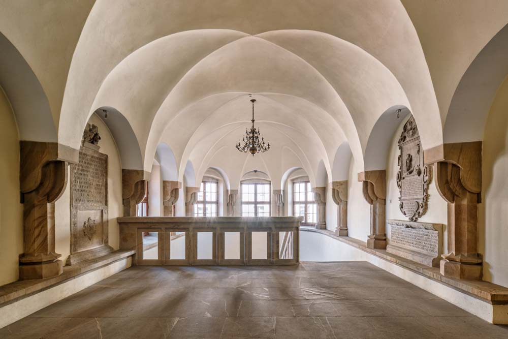 Wnętrze Ratusza w Lwówku Śląskim z kamiennymi łukami i sklepieniem krzyżowym. Na ścianach widoczne są epitafia i tablice pamiątkowe, a przestrzeń oświetla żyrandol.
