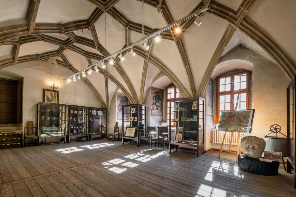 Wnętrze Ratusza w Lwówku Śląskim z gotyckim sklepieniem żebrowym. W sali ustawione są gabloty z eksponatami muzealnymi i planszami informacyjnymi.