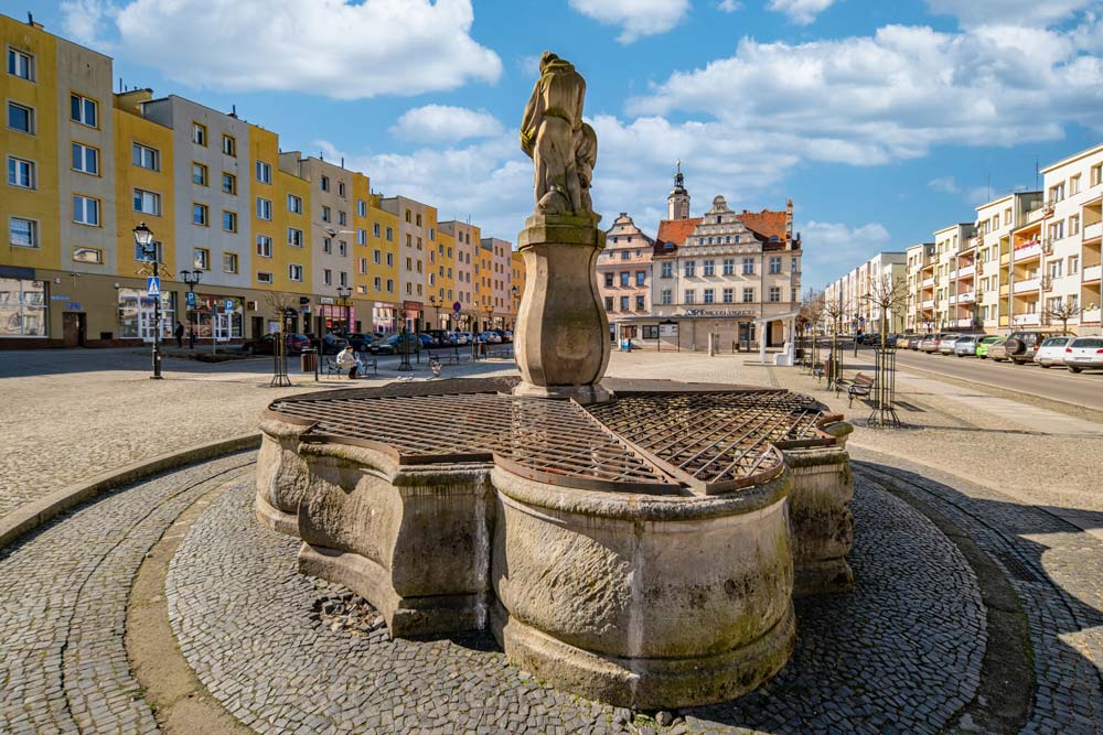 Kamienna fontanna na lwóweckim rynku z rzeźbą w centrum. W tle widoczne są kolorowe kamienice i wieża ratusza.