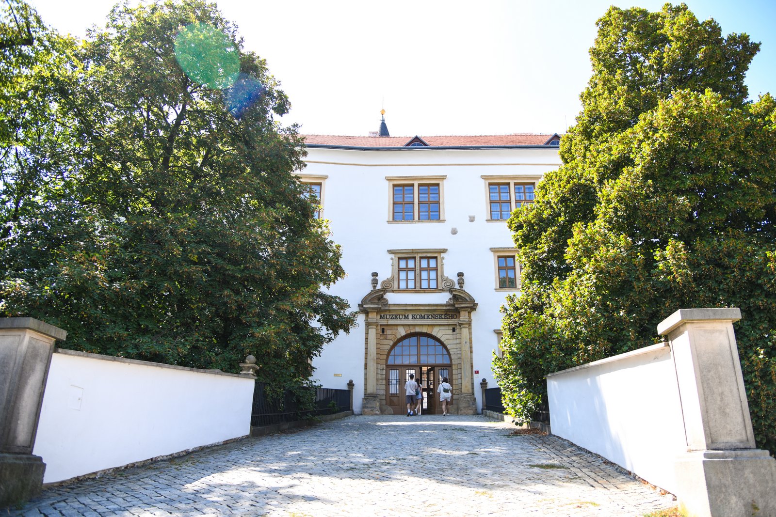 Wejście do Muzeum Jana Amosa Komenského w Přerovie w pałacu o białej elewacji, otoczonym drzewami, z napisem nad portalem wejściowym.