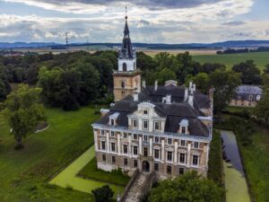 Ujęcie z lotu ptaka na Zamek Roztoka, otoczony fosą i parkiem. Widać rozległy pałacowy kompleks z centralną wieżą i panoramę pól oraz lasów w tle.