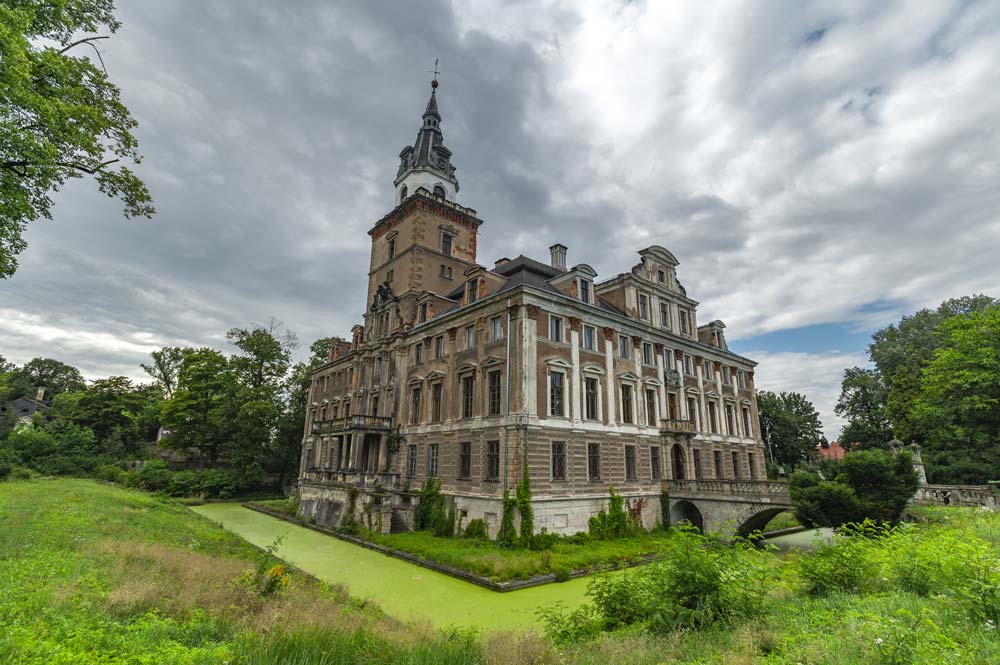 Zamek Roztoka otoczony zieloną fosą, z barokową fasadą i wysoką wieżą. Budynek ma bogato zdobione elewacje i znajduje się w otoczeniu parku.