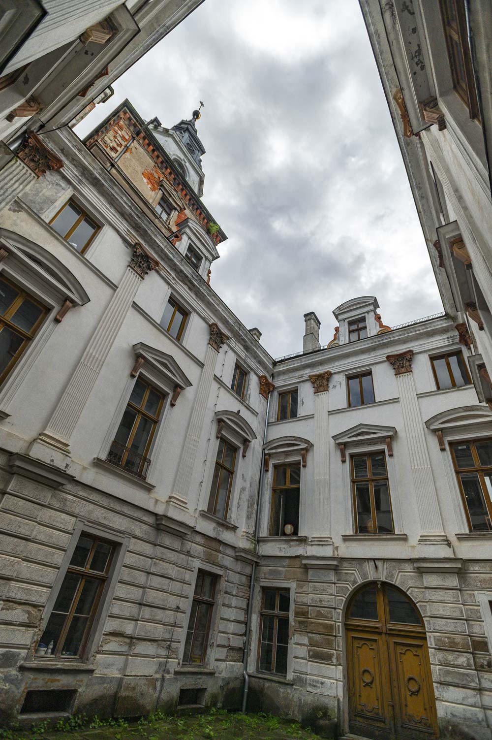 Widok z dziedzińca Zamku Roztoka na otaczające go elewacje z wysokimi oknami i kolumnami. Na górze widać wieżę z ozdobnym hełmem, a nad budynkiem wiszą szare chmury.