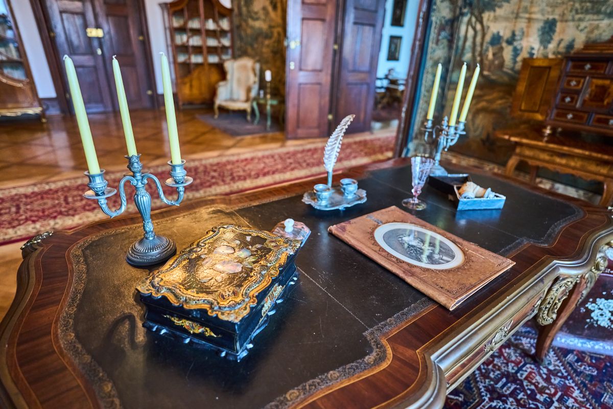 Zbliżenie na eleganckie biurko w Zamku w Náchodzie z ozdobnym kałamarzem, świecznikiem i szkatułką. W tle widać fragment biblioteczki i tapiserię ścienną.