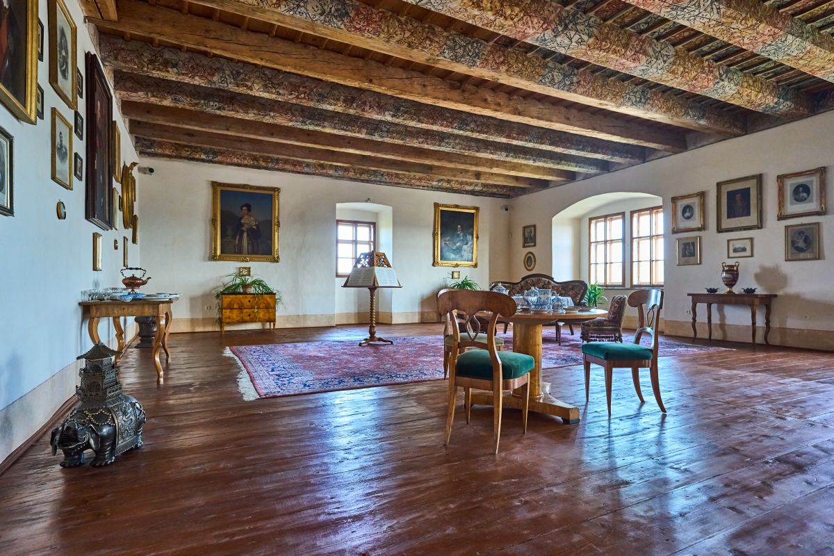 Przestronny pokój w Zamku w Náchodzie z malowanym belkowanym stropem, drewnianą podłogą i zabytkowymi meblami. Ściany zdobią portrety i klasyczne dekoracje.