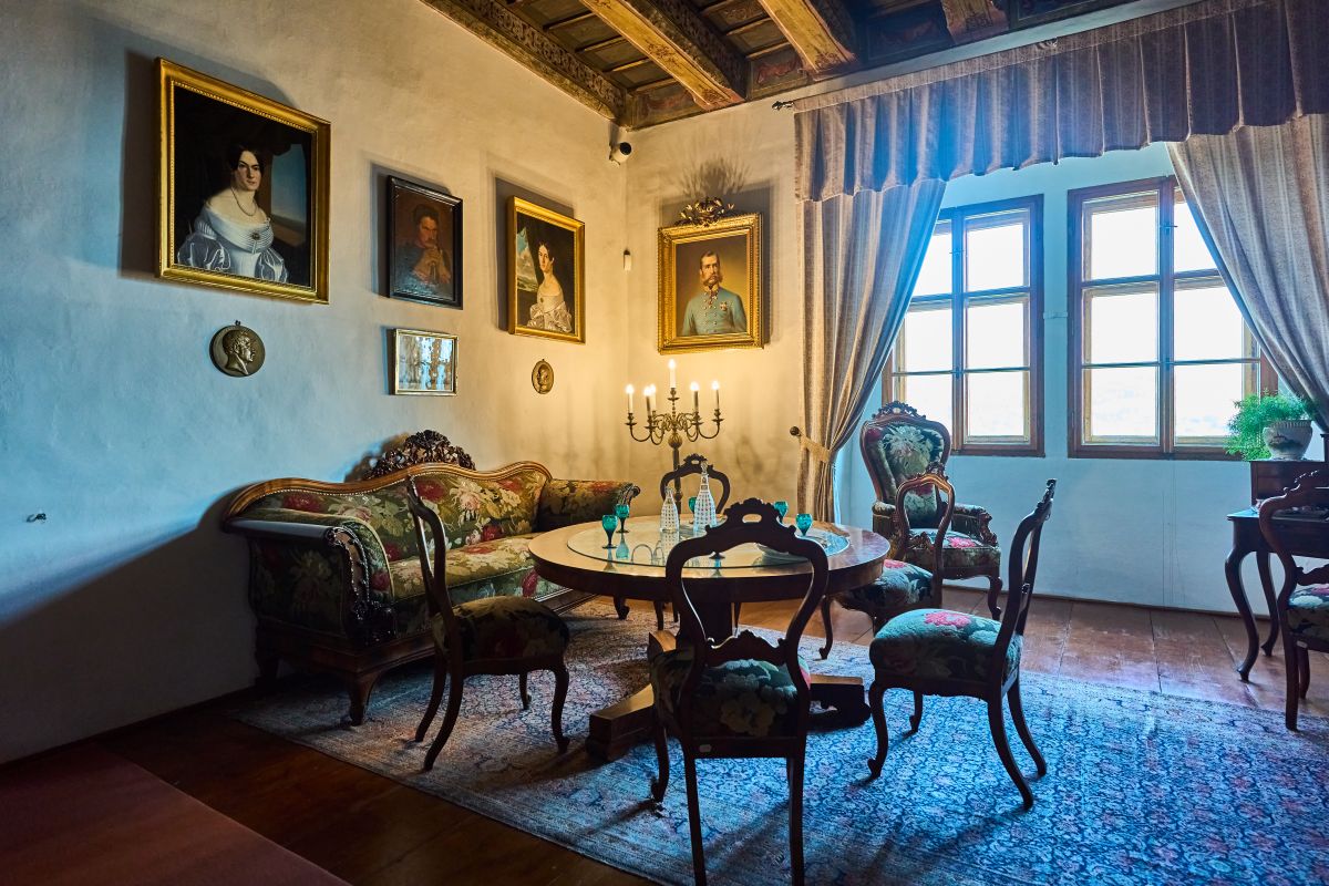 Stylowy salon w Zamku w Náchodzie z drewnianym sufitem i meblami tapicerowanymi kwiecistym materiałem. Na ścianach wiszą portrety arystokratów, a w centrum stoi okrągły stół z kieliszkami.