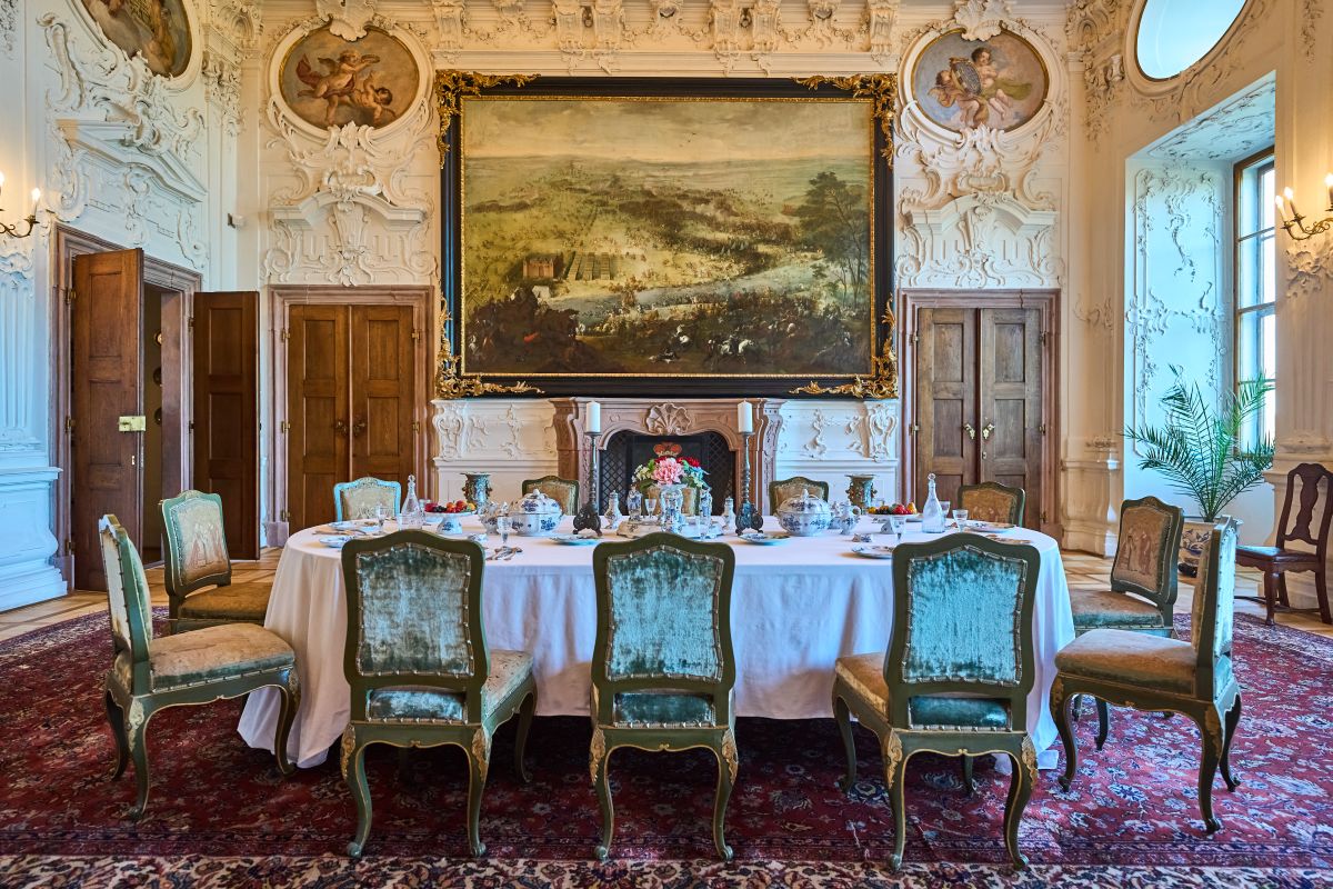 Reprezentacyjna jadalnia w Zamku w Náchodzie z długim stołem nakrytym porcelaną i szkłem. Ściany zdobią sztukaterie i ogromny obraz nad kominkiem w złotej ramie.