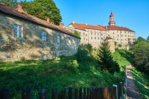 Panorama wzgórza z Zamkiem w Náchodzie, otoczonym murami obronnymi i zielenią. Widać kamienne zabudowania z czerwonym dachem i wieżę z charakterystyczną czerwoną kopułą.